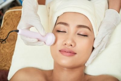 Close-up shot of beautician hands making ultrasound facial procedure while pretty Asian client lying on treatment table with closed eyes